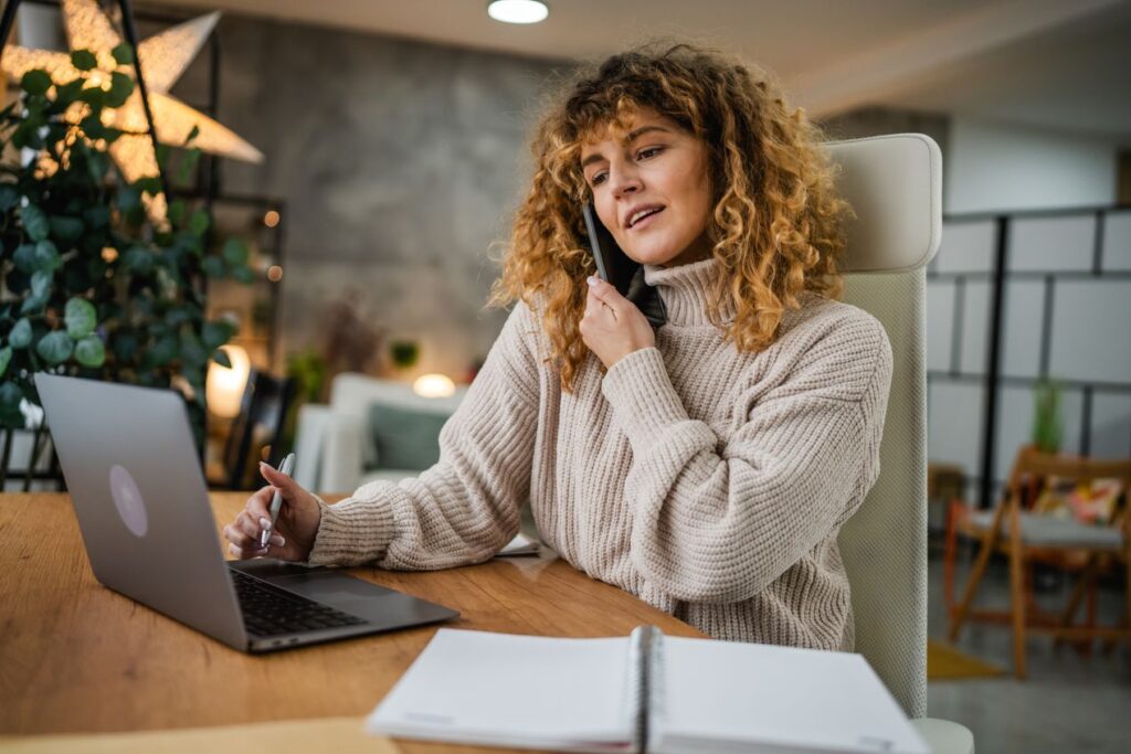 Frau mit Locken arbeitet im Homeoffice an einem Laptop und telefoniert, während sie Inhalte für E-learning Content Produktion überprüft.