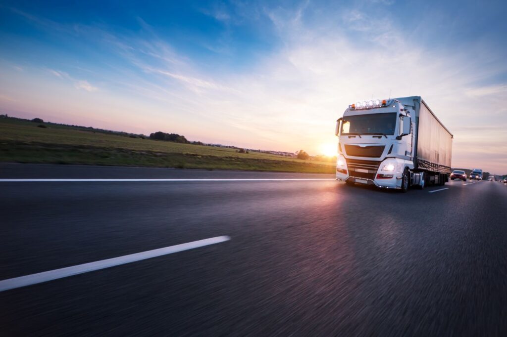 Moderner LKW mit eingeschalteter Beleuchtung fährt bei Sonnenuntergang über eine Landstraße.