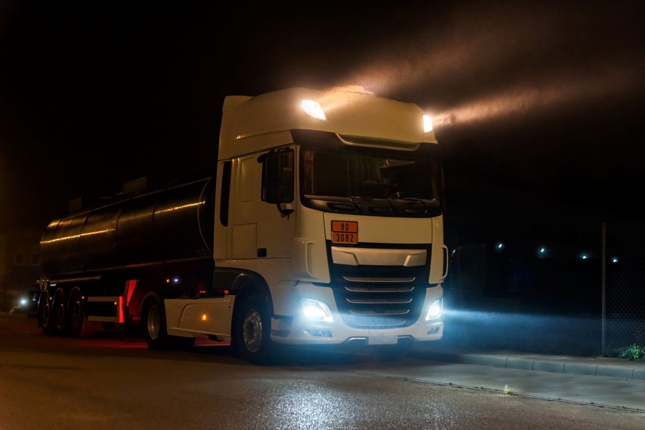 Ein weißer Tanklastwagen mit eingeschalteter Beleuchtung steht nachts auf einem Parkplatz.